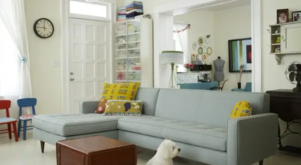 Un salotto accogliente con divano azzurro, cuscini decorati e un cane bianco. L'ambiente è dominato da **colori pastello** e dettagli vintage.