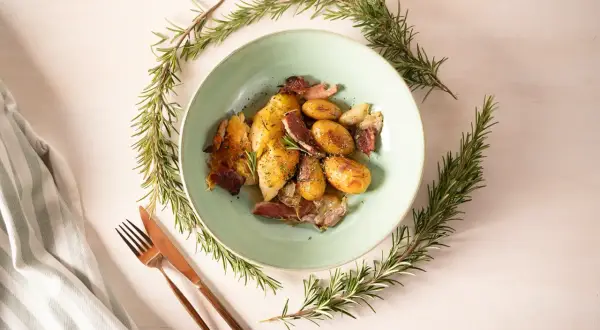Ein Teller mit Hähnchen, Kartoffeln und Speck, umrahmt von Rosmarinzweigen. Die Rosmarin verwendung verleiht dem Gericht ein wunderbares Aroma.