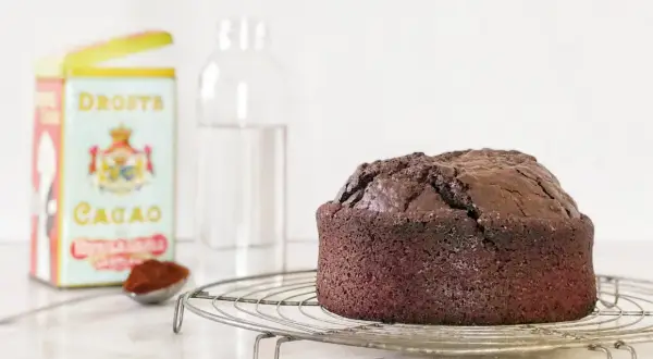 Torta all'acqua al cacao appena sfornata, su una gratella. Accanto, una scatola di cacao Droste e un bicchiere d'acqua.