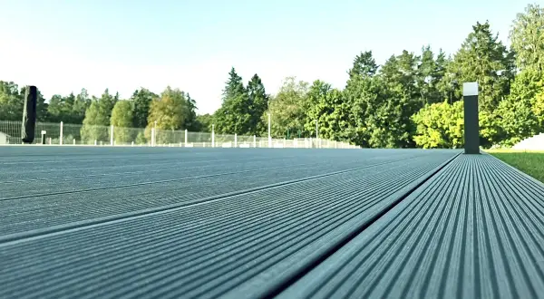 Szara deska kompozytowa tworzy nowoczesną nawierzchnię tarasu, otoczoną zielenią i jasnym niebem.