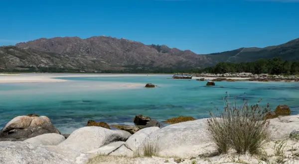Aguas turquesas y arena blanca bordean la costa, con el imponente monte Pindo como telón de fondo. Rocas cubiertas de musgo salpican la orilla.