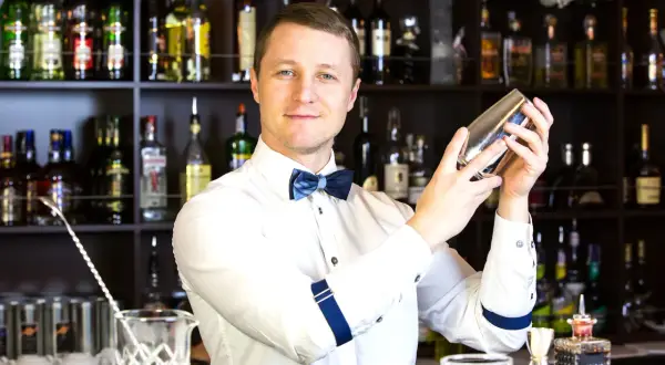 Uśmiechnięty barman w białej koszuli i niebieskiej muszce miesza drinka w shakerze na tle regałów z butelkami alkoholu.
