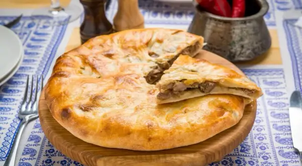 Chaczapuri co to? Złocisty placek z mięsnym farszem, podany na drewnianej desce, z papryczkami chili w tle. Pyszne danie gruzińskie.