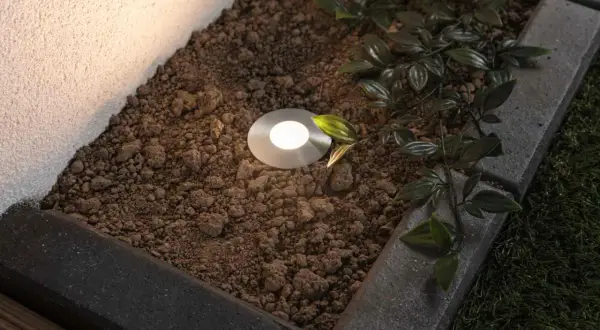 Oświetlona okrągła lampa dogruntowa w ziemi, otoczona kamieniami i zielonymi liśćmi.