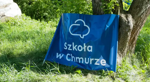 Niebieski baner z napisem "Szkoła w Chmurze" oparty o drzewo, na tle zielonej trawy i liści.