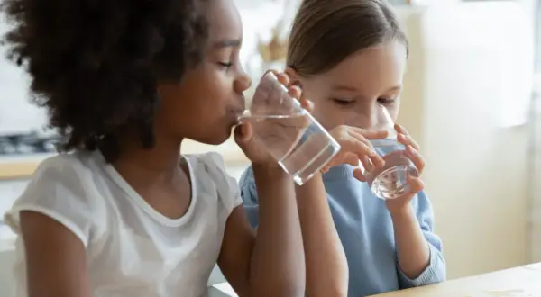 Deux enfants, une fille noire et une fille blanche, boivent de l'eau à la même table. L'eau est essentielle pour chaque enfant.