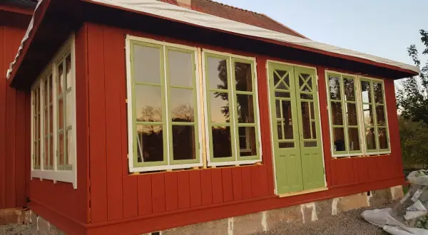Ett rött hus med pågående arbete för att bygga glasveranda. Gröna fönster och dörrar reflekterar himlen.