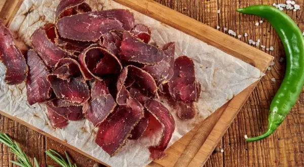 Suszona wołowina przepis na pyszne przekąski. Na desce ułożone cienkie plastry mięsa, obok zielona papryczka chili i rozmaryn.