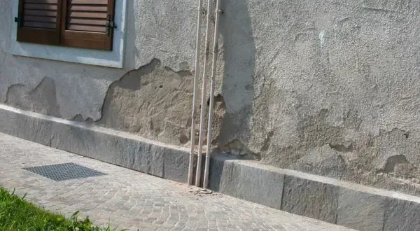 Il muro esterno grezzo mostra scrostamenti e necessita di essere pitturato. Tre tubi bianchi corrono verticalmente lungo la parete.
