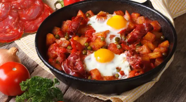Pyszny obiad z jajek, ziemniaków, chorizo i groszku w patelni. Obok pomidory, cebula i natka pietruszki.