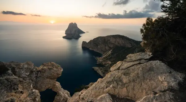 Vista del ojo de es Vedrà al atardecer, con el sol poniente tiñendo el cielo de naranja y el mar en calma.
