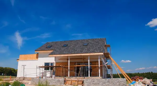 Budowa domu 120m2 w toku. Białe ściany, szary dach, rusztowania. W tle błękitne niebo i zieleń.