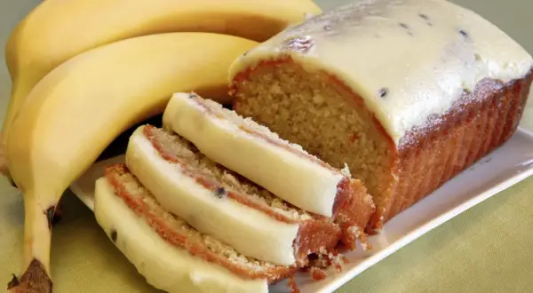 Wilgotne ciasto bananowe z kremową polewą i kawałkami owoców, obok dojrzałych bananów.