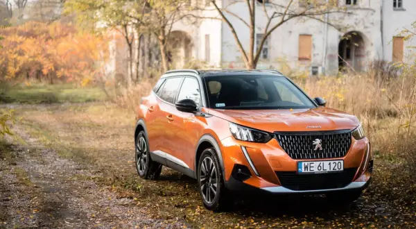 Pomarańczowy Peugeot 2008 GT na tle jesiennego krajobrazu z opuszczonym budynkiem.