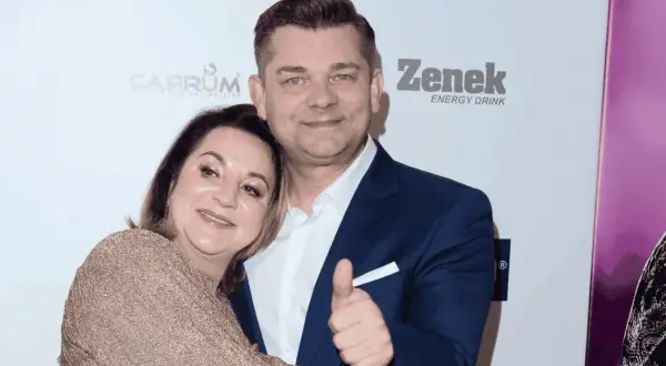 Danuta Martyniuk, w złotej sukience, przytula się do mężczyzny w granatowym garniturze, który pokazuje kciuk w górę.