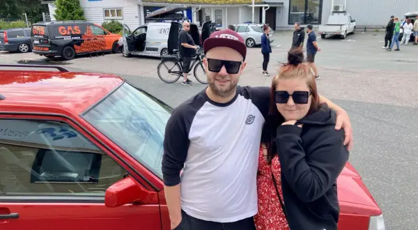 Ett par poserar framför en röd bil, redo att bygga ljud i bil. I bakgrunden syns bilar och folk vid en butik.