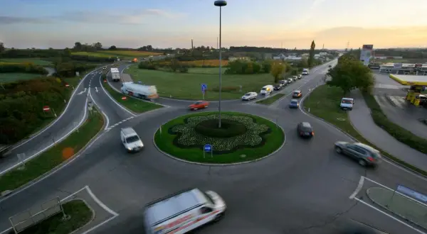 Ruchliwe rondo z wieloma rodzajami rond, samochodami i ciężarówkami. W tle widać pola i niebo o zachodzie słońca.