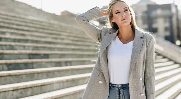 Chiara Ferragni w szarej marynarce w kratę i jeansach, z ręką we włosach, na tle schodów.