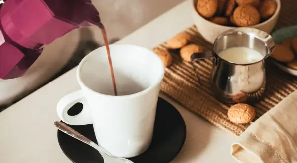 Un buon caffè fumante viene versato da una moka viola in una tazza bianca, accompagnato da biscotti e latte.