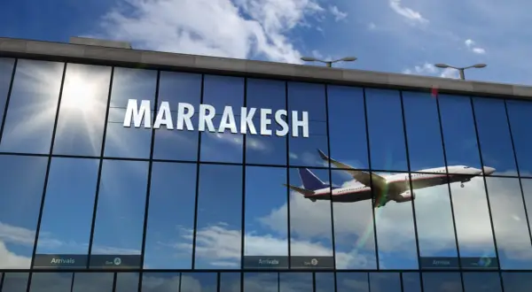 Samolot wznosi się na tle błękitnego nieba z chmurami, odbijając się w szklanej fasadzie lotniska w Marrakeszu.
