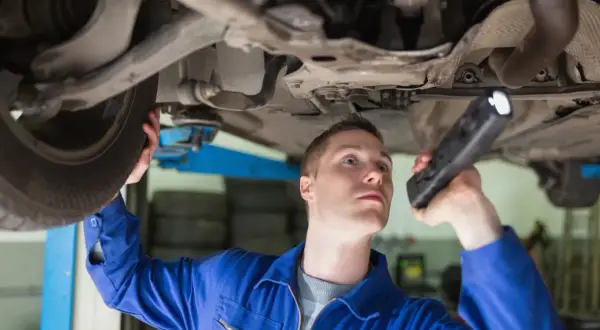 Mechanik w niebieskim kombinezonie bada podwozie auta latarką, przygotowując się do wymiany sworznia wahacza.