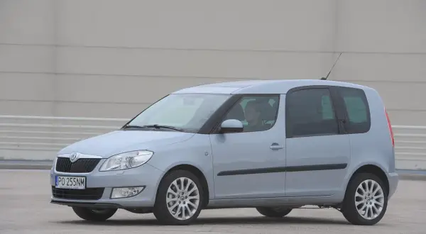 Jasnoszary Skoda Roomster z kierowcą za kierownicą stoi na betonowym placu przed szarym budynkiem.
