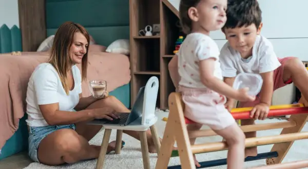Kobieta pracuje na laptopie, pijąc kawę, podczas gdy dzieci bawią się obok. Pokazuje, jak pogodzić pracę z opieką nad dzieckiem.