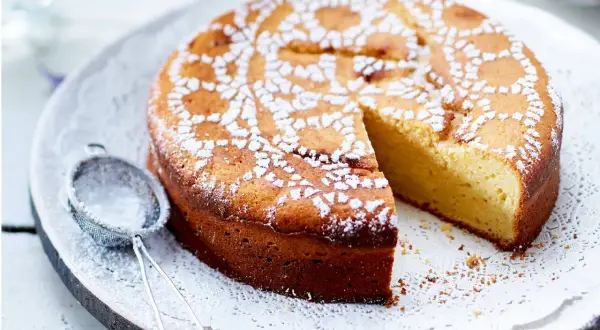 Ein saftiger Joghurt Kuchen, mit Puderzucker bestäubt und einem Stück herausgeschnitten. Daneben liegt ein kleines Sieb.