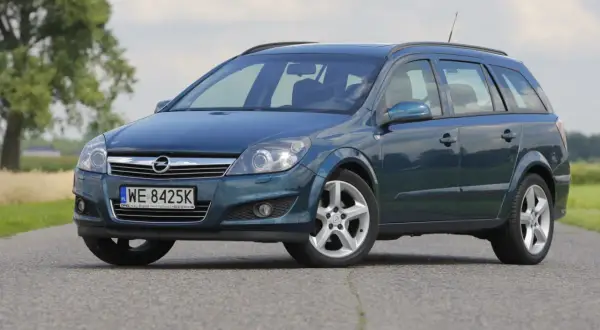 Granatowy Opel Astra Cosmo w wersji kombi stoi na asfaltowej drodze, otoczony zielenią i drzewami.