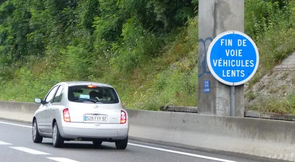 Voiture argentée sur l'autoroute, passant devant un panneau "Fin de voie véhicules lents". La nature luxuriante borde la route.
