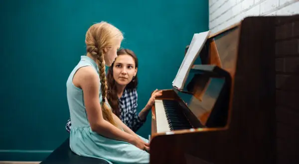 Dziewczynka z warkoczem uczy się gry na pianinie pod okiem nauczycielki. Nuty na pulpicie wskazują na lekcję muzyki.