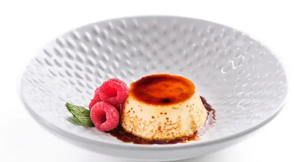 Delicioso flan de queso fresco con caramelo líquido, decorado con frambuesas y menta en un plato blanco texturizado.