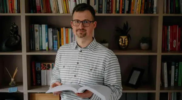 Mężczyzna w okularach, trzymający otwartą książkę, z neutralnym wyrazem twarzy, jakby uczył się, jak nie okazywać emocji na twarzy.