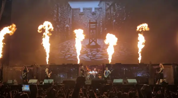 Płomienie buchają na scenie, gdzie grają zespoły rockowe. W tle widać zamek i gilotynę.