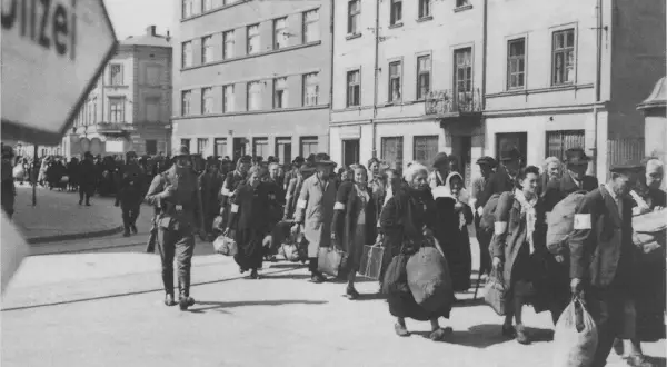 Människor med armbindlar ledsas förbi Schindlers fabrik. En soldat går framför.