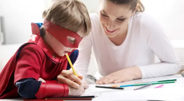 Chłopiec w stroju superbohatera rysuje, a mama z uśmiechem wspiera go, pokazując, jak zachęcić dziecko do nauki przez zabawę.
