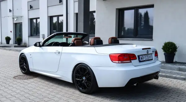 Białe BMW E93 cabrio z brązową skórzaną tapicerką i czarnymi felgami stoi na kostce brukowej przed nowoczesnym budynkiem.