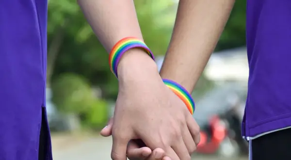 Zwei Hände halten sich, geschmückt mit Regenbogenarmbändern. Eine Person trägt ein lila Oberteil. Ein Symbol für Liebe, das halb Mann halb Frau sein kann.