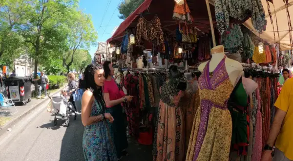 Persone curiosano tra le bancarelle colorate della fiera di San Ciriaco, ammirando abiti etnici e tessuti vivaci sotto il sole.