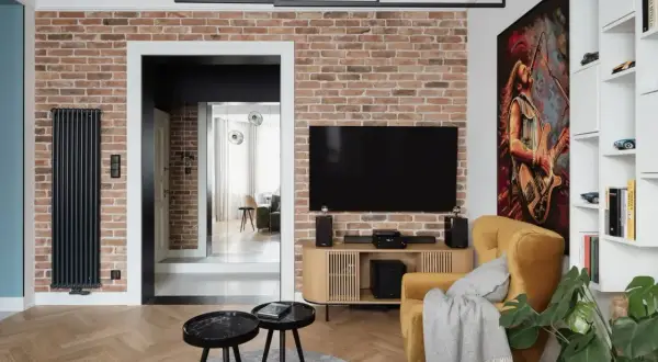 Nowoczesny salon z ceglaną ścianą, na której subtelne oświetlenie kamienia podkreśla jego fakturę. Obok telewizor i żółty fotel.