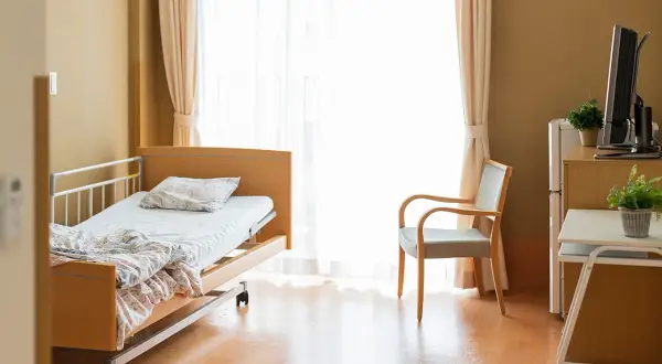 Ein helles Zimmer mit einem Pflegebett auf Rezept AOK, einem Sessel und einem Fernseher.
