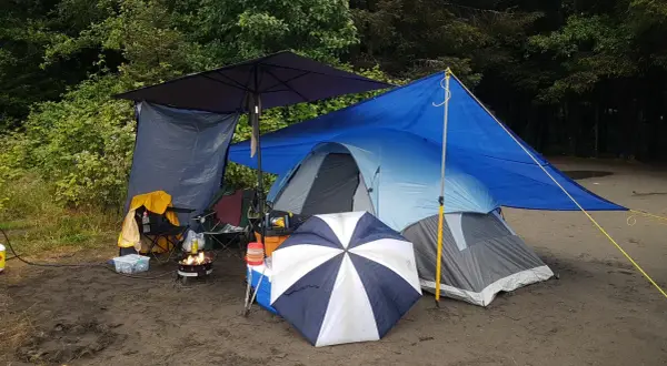 Campingplats med tält, eldstad och paraply. Ved torkar under presenning.