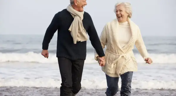 Szczęśliwa para seniorów biegnie plażą, trzymając się za ręce. Idealne tanie wycieczki dla seniorów, by cieszyć się wspólnym czasem.