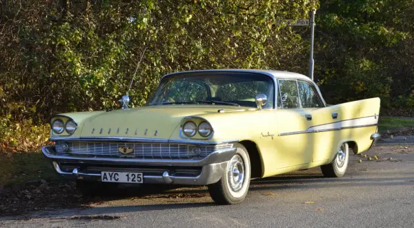 Gul Chrysler Saratoga från 1950-talet, med kromdetaljer och vita däcksidor, parkerad vid en lummig buske.