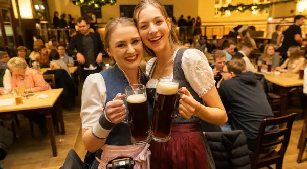 Zwei fröhliche Frauen in Tracht halten je ein großes, gestauchtes Bier. Im Hintergrund ist eine belebte Gaststube zu sehen.