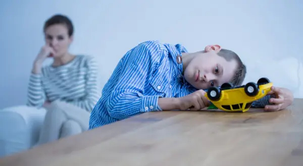 Chłopiec z zespołem Aspergera bawi się zabawką, podczas gdy matka w tle wygląda na zmartwioną, być może zmagając się z jego agresją.
