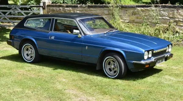 Blå Reliant Scimitar GTE, en klassisk sportsvagn med kombikaross, parkerad på en gräsmatta framför en stenmur och en grind.