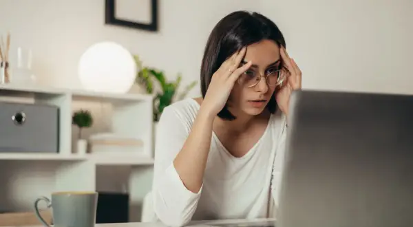 Zestresowana kobieta z okularami masuje skronie, patrząc na ekran laptopa. Brak odpowiedzi na reklamację pogłębia jej frustrację.
