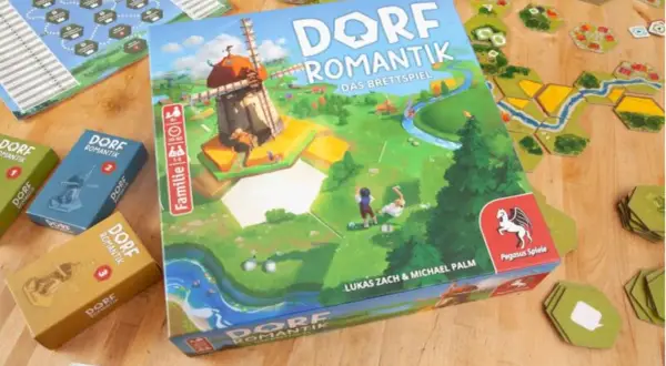 Das Familienspiel des Jahres "Dorfromantik" mit seinen bunten Spielsteinen und der Hauptbox, die eine idyllische Landschaft mit Windmühle zeigt.