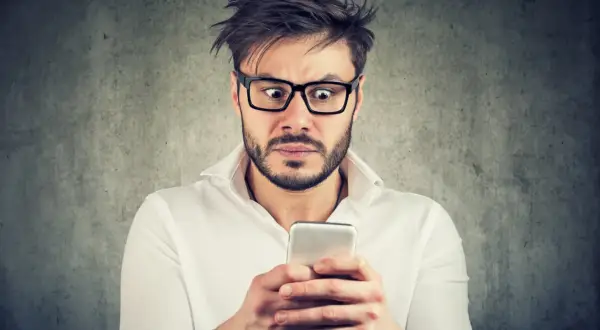 Przerażony mężczyzna w okularach czyta na telefonie o tym, co grozi za oczernianie drugiej osoby.